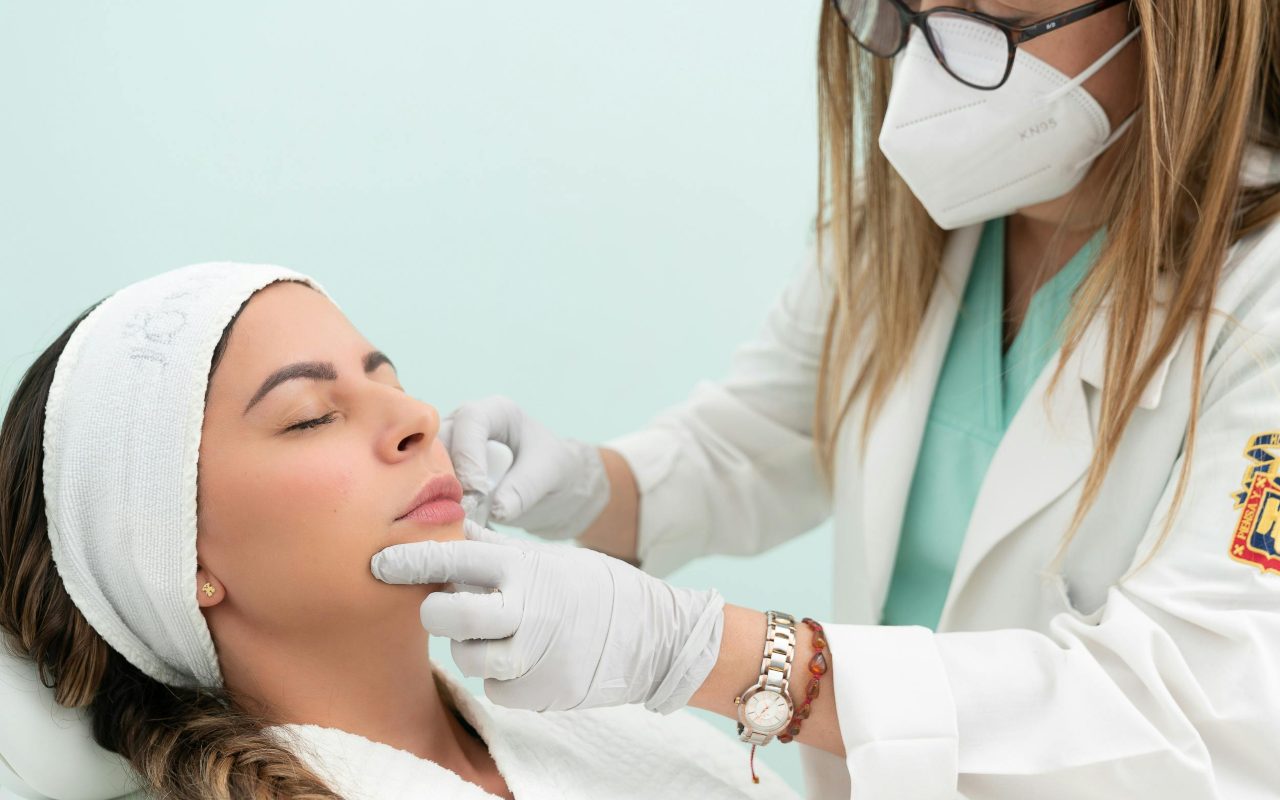 Deevojka leži na krevetu dok joj doktorka ubrizgava špric u lice.