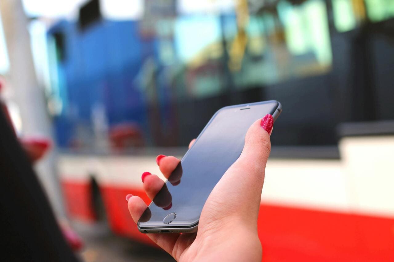 Ruka sa crvenim lakiranim noktima drži crni pametni telefon ispred autobusa